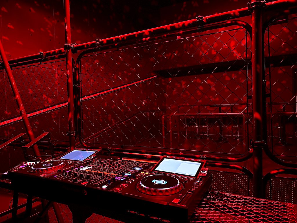 The DJ booth of 3F SECRET CLUB: DJ equipment inside a wire-mesh enclosure under red light