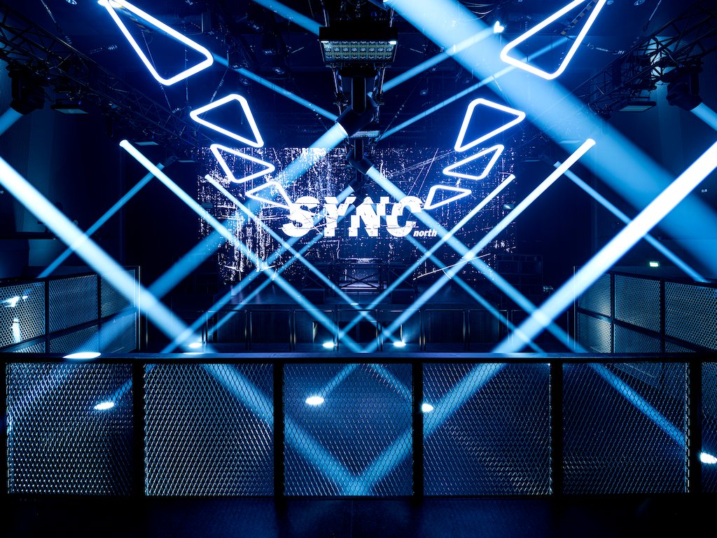 Blue beams crossing above the main floor, with the SYNC north LED sign behind