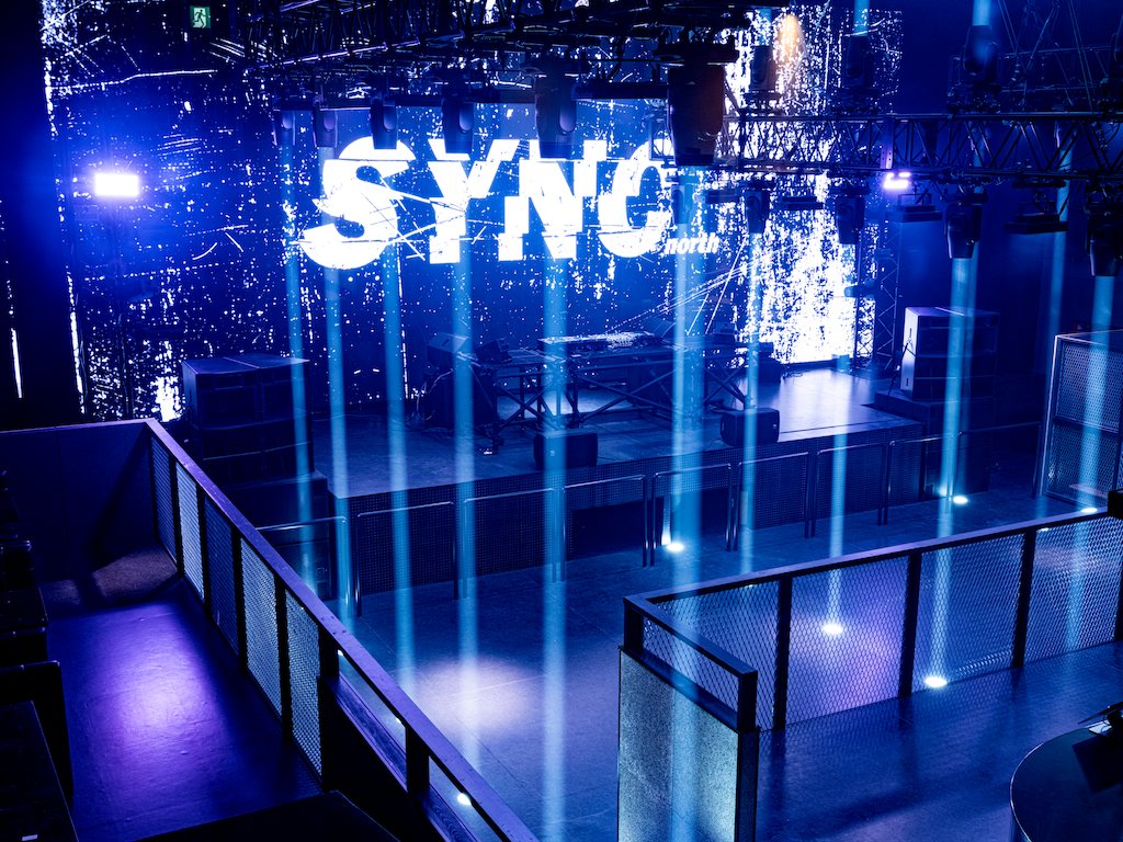 The whole floor seen over the upper railing: columns of blue light and the SYNC logo behind