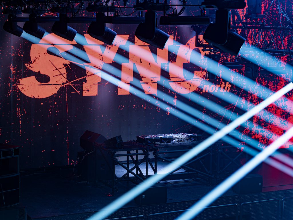 Pale blue laser beams crossing in front of the SYNC north logo on the LED wall