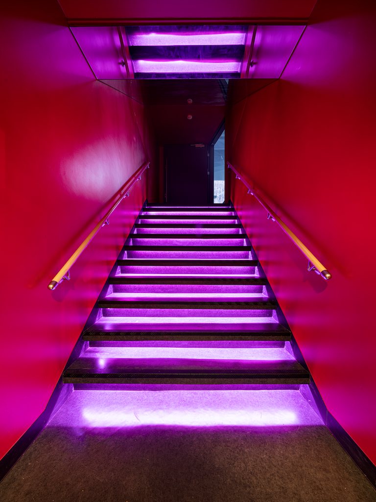 An interior stairway with glowing step edges, handrails on both sides and pink-lit walls